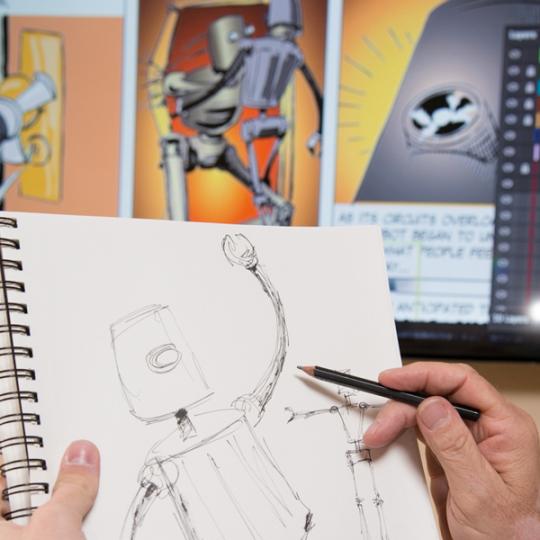 A student is working on a drawing of a robot, holding a pencil and sketch pad up to a computer to compare their graphic design to a completed and colored illustration.