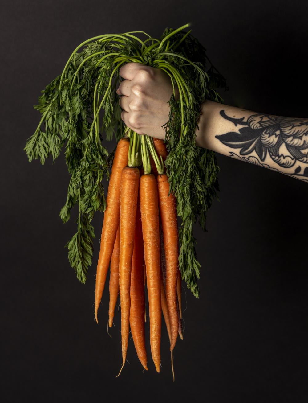 Photo of tattooed arm holding a bunch of carrots by the stems - photo by Mercedes Gania