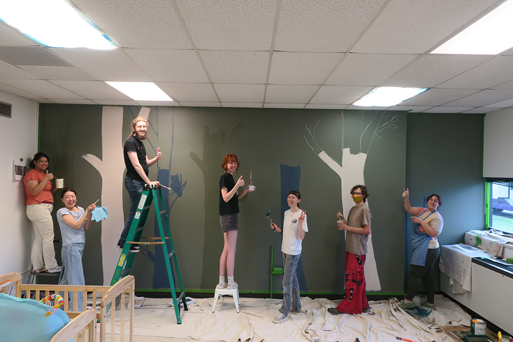 ACC Studio Art Capstone students painting a mural of trees and animals in ACC's Child Development Center, giving thumbs up to the camera.