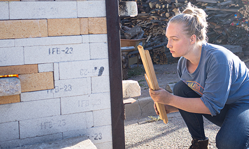 ACC student standing near kiln with wood in her hands.