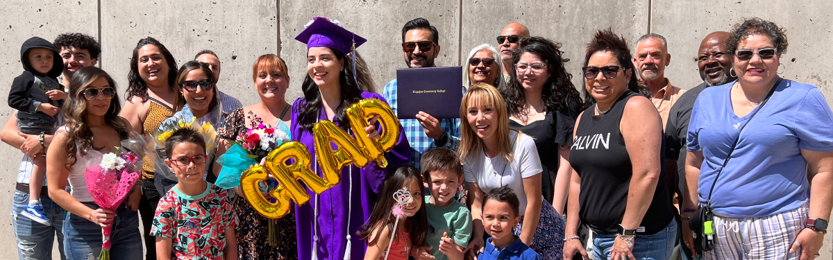 ACC graduate in regalia holding GRAD balloon and surrounded by friends/family outside of the main building at the Littleton Campus.