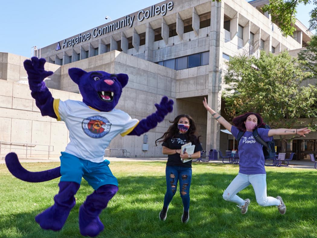 ACC Puma jumping in the air with 2 students outside of the main building at the Littleton Campus.