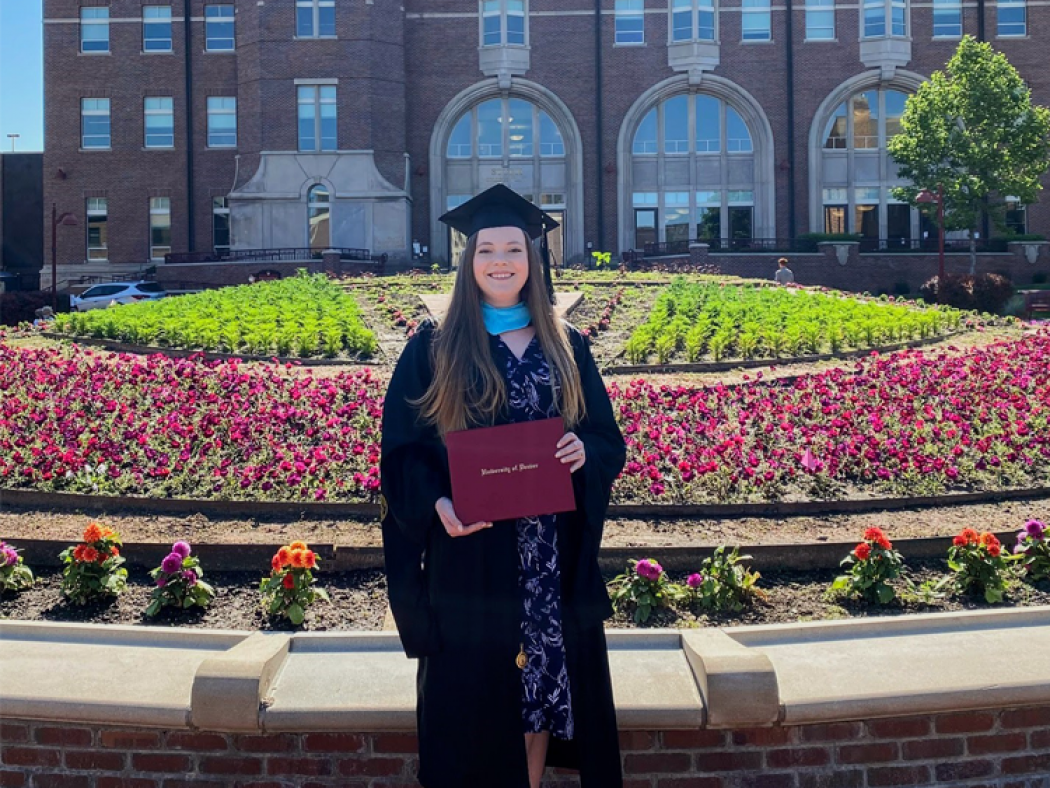 Amanda Savarese in graduation regalia in front of flowers and college building.