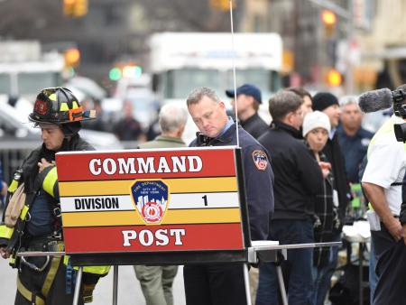 A firefighter and a police officer manage a crisis at a mobile command post. Members of the public and emergency vehicles behind them.