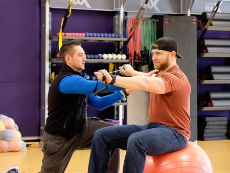 A physical therapist in the ACC gym assists a man on a yoga ball, adjusting his grip on a training band.