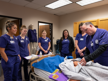 ACC Nursing students in classroom at Littleton Campus with instructor showing procedures with manikin on gurney. 