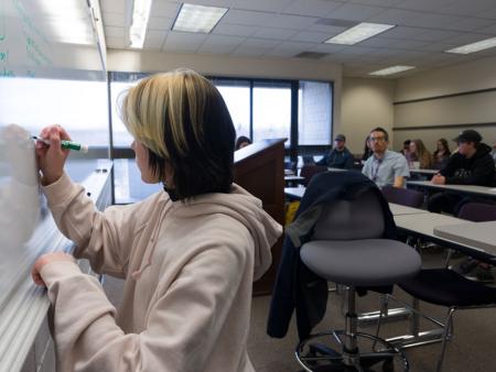 A student writes on a white board with her classroom behind her.