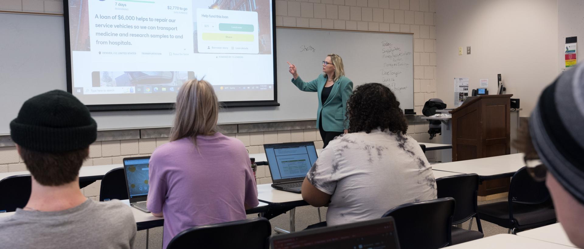 ACC Economics faculty member in front of classroom, discussing points on a screen at the front of a Littleton Campus classroom.