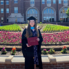 Amanda Savarese in graduation regalia in front of flowers and college building.